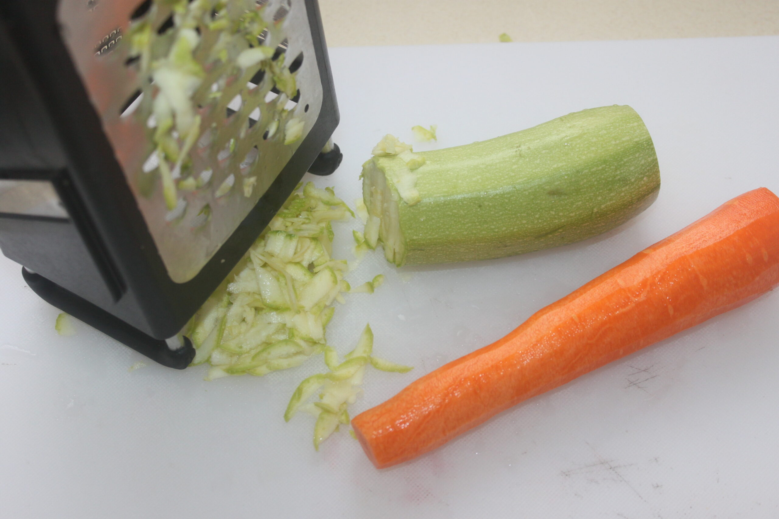 Une râpe à boîte avec des courgettes râpées, une courgette partiellement râpée et une carotte entière se trouvent sur une planche à découper blanche, prêtes pour votre prochaine recette de Brouillon automatique.
