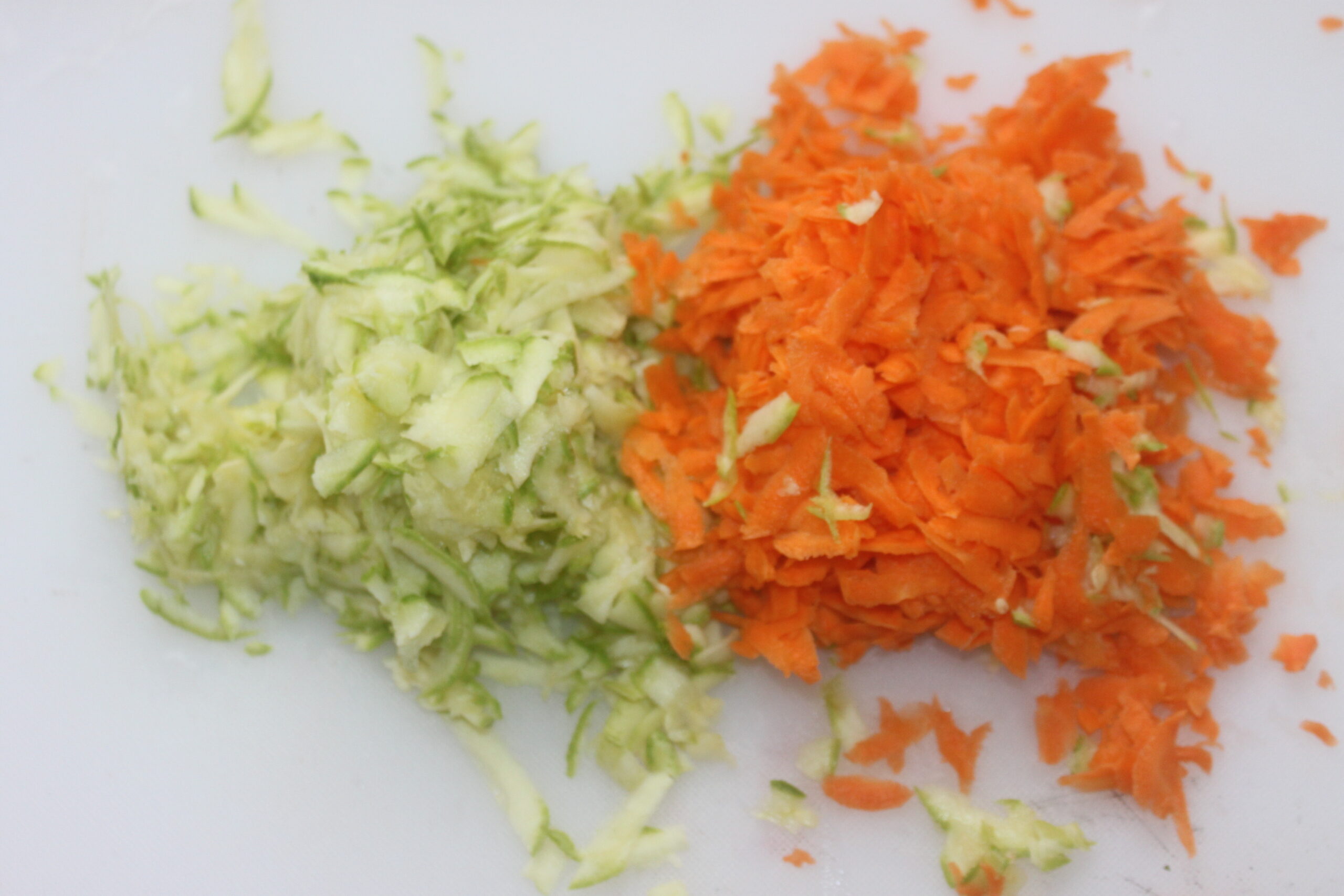 Des courgettes et des carottes finement râpées sont placées côte à côte sur une planche à découper blanche, prêtes pour votre prochaine recette de Brouillon automatique.