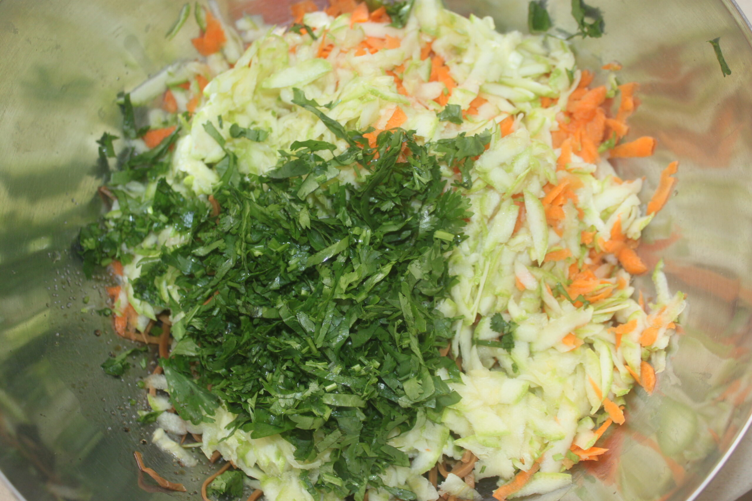 Un bol en métal contenant des courgettes râpées, des carottes râpées et de la coriandre fraîche hachée est prêt pour votre prochaine recette auto inspirée de Brouillon.