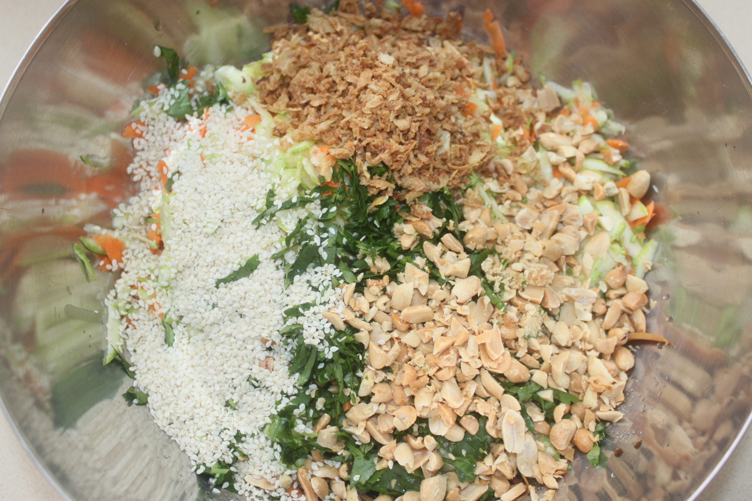 Un bol en métal contenant des cacahuètes hachées, des graines de sésame, des échalotes frites, des légumes râpés et des herbes, séparés en sections, parfait pour ajouter à vos plats auto Brouillon préférés.