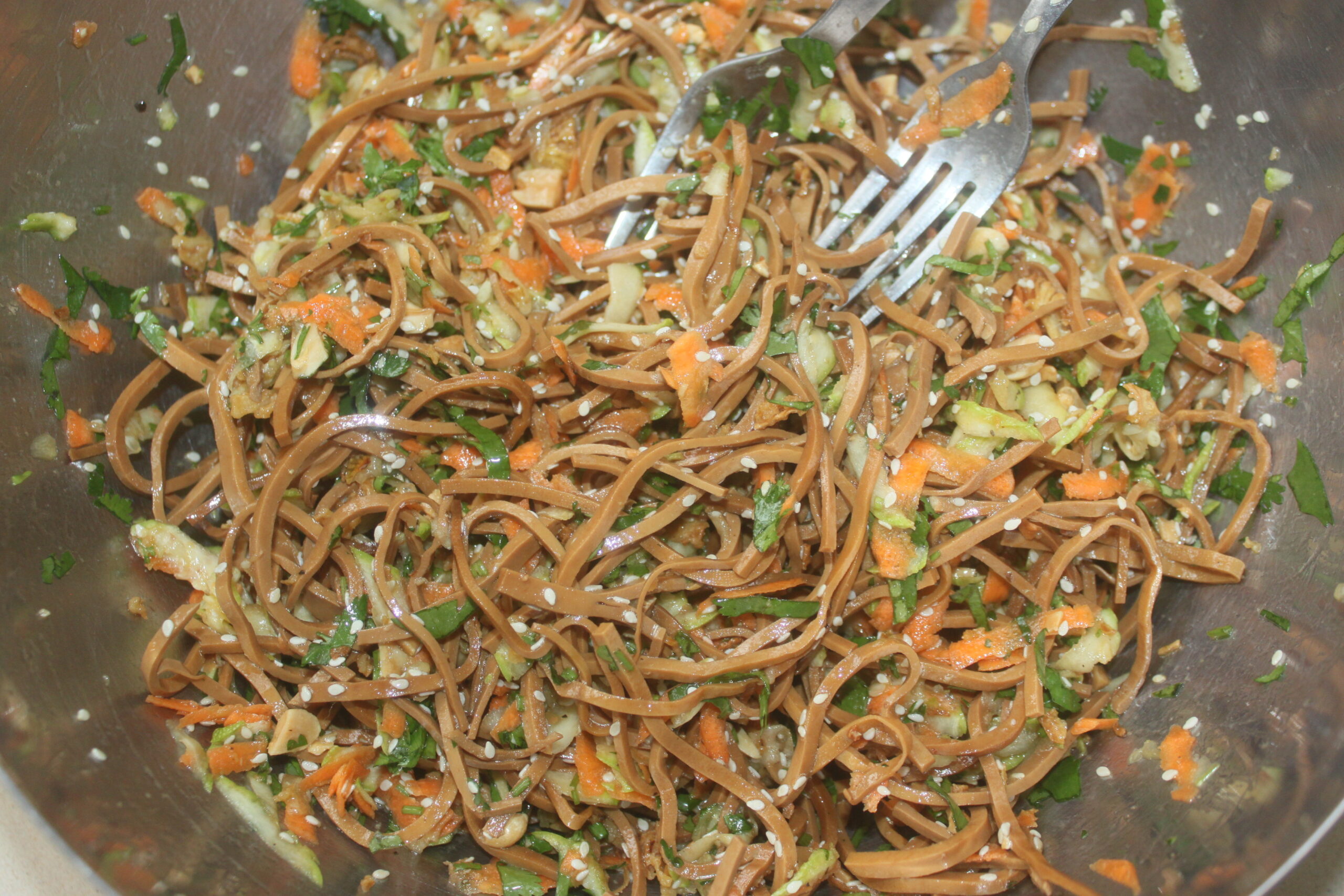 Un bol en métal rempli de salade de nouilles soba, mélangée à des carottes râpées, des courgettes, des légumes verts, des graines de sésame et deux fourchettes, parfait pour un déjeuner rapide en auto Brouillon.