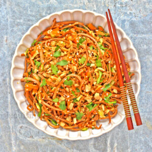 Un bol de nouilles sautées garnies d'arachides hachées et d'herbes, inspiré de la salade végétarienne, servi avec des baguettes en bois sur une surface grise texturée.