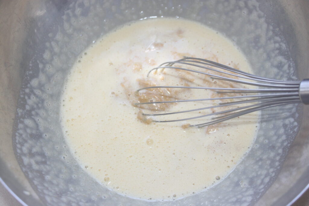 Un fouet en métal mélange une pâte à cake banane marron clair dans un grand bol en métal.