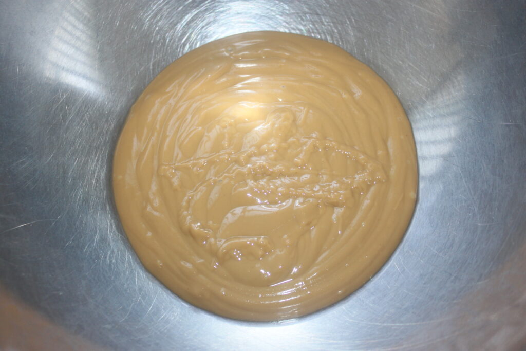 Une mare de liquide épais et brun clair, probablement du lait concentré sucré, s'est étalée au fond d'un bol à mélanger en métal, prête à devenir la base riche d'un délicieux Cake banane.