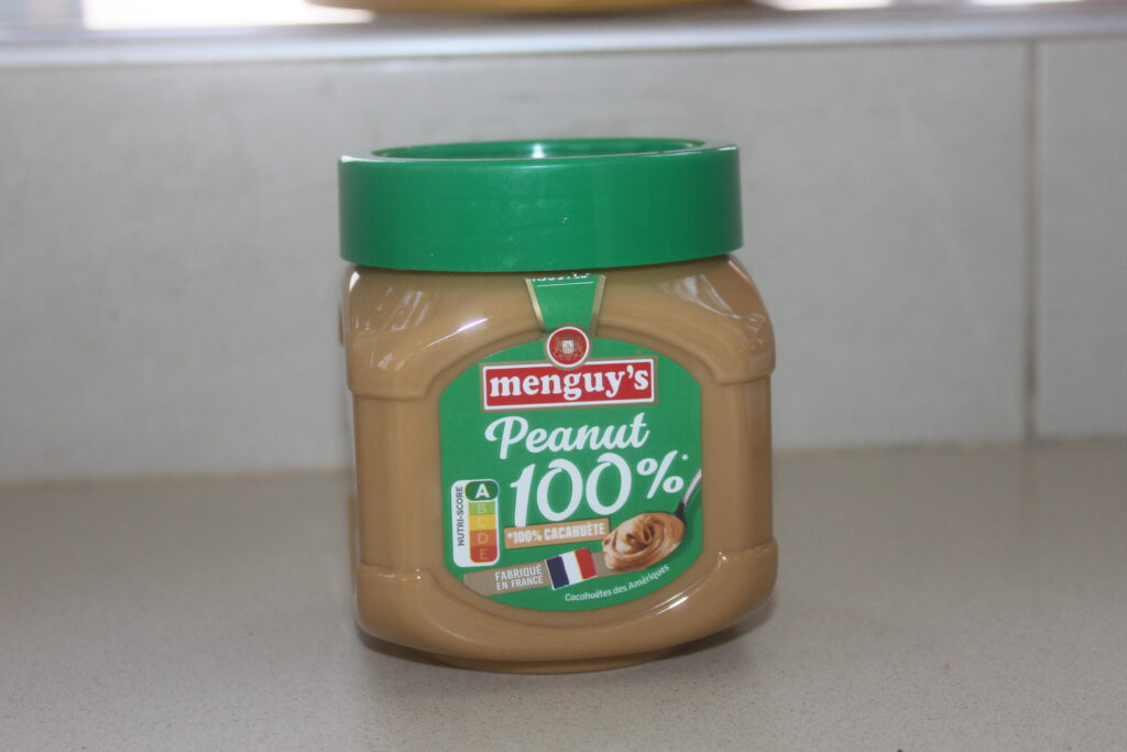 Un pot de beurre 100% cacahuète de Menguy au couvercle vert trône sur le comptoir d'une cuisine, parfait pour agrémenter les tartines du matin ou une délicieuse galette de banane.